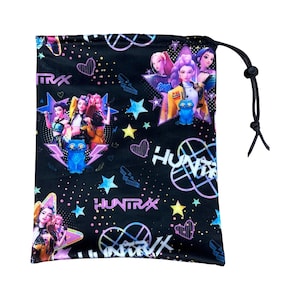 以下が含まれることがあります： 「HUNTRAX」の文字が入ったカラフルなグラフィックデザインの黒い巾着袋。デザインには、アニメのキャラクター、星、ハート、幾何学模様がピンク、青、黄色、紫の色合いで描かれています。バッグは黒い巾着開閉式です。