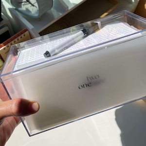 May include: A clear plastic box with a white interior and the words "one two" printed on the front. The box is filled with small white cards and a white marker.