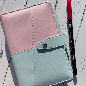 May include: A light blue and pink leather planner with a silver snap closure. A black marker with a red cap and the text "7.55 Tombow ABT" is next to the planner.