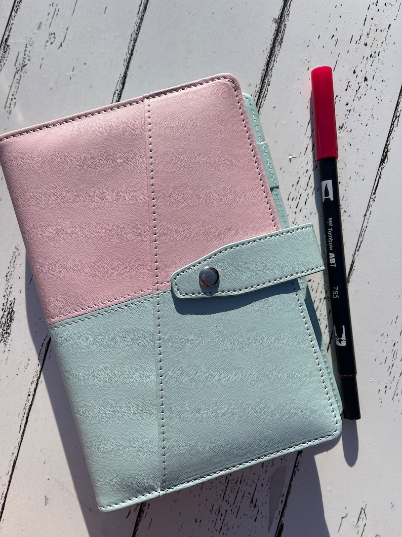 May include: A light blue and pink leather planner with a silver snap closure. A black marker with a red cap and the text "7.55 Tombow ABT" is next to the planner.