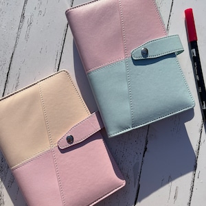 May include: Two pastel-colored leather bound journals with a flap closure and a silver button. The journals are on a white wooden surface.