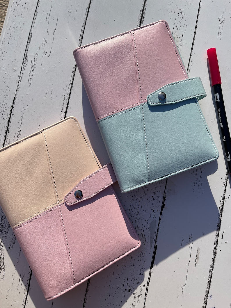 May include: Two pastel-colored leather bound journals with a flap closure and a silver button. The journals are on a white wooden surface.