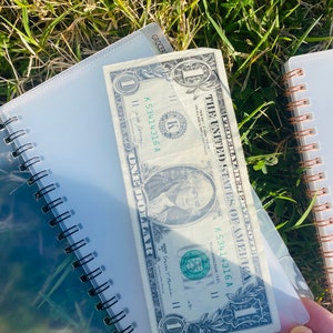 Peut inclure: Un billet d'un dollar avec le num&eacute;ro de s&eacute;rie K53414316 A est plac&eacute; dans une pochette en plastique transparente. Le billet est pos&eacute; sur un carnet &agrave; spirales en plastique transparent.