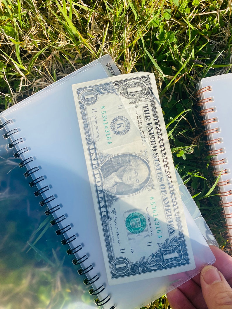 Peut inclure: Un billet d'un dollar avec le num&eacute;ro de s&eacute;rie K53414316 A est plac&eacute; dans une pochette en plastique transparente. Le billet est pos&eacute; sur un carnet &agrave; spirales en plastique transparent.