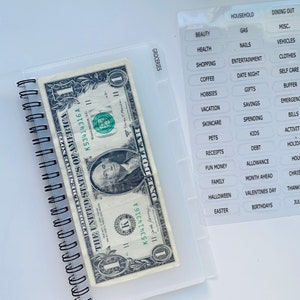 Peut inclure: Un classeur en plastique transparent contenant un billet d'un dollar. Le classeur est dot&eacute; d'une fermeture &agrave; glissi&egrave;re et d'une poche transparente sur le devant. La poche contient une vari&eacute;t&eacute; d'&eacute;tiquettes pour diff&eacute;rentes cat&eacute;gories, telles que "&eacute;picerie", "m&eacute;nage", "repas au restaurant", "beaut&eacute;", "essence", "divers", "sant&eacute;", "ongles", "v&eacute;hicules", "achats", "divertissements", "v&ecirc;tements", "caf&eacute;", "soir&eacute;e", "soins personnels", "loisirs", "cadeaux", "tampon", "vacances", "&eacute;pargne", "urgences", "soins de la peau", "d&eacute;penses", "factures", "animaux de compagnie", "enfants", "activit&eacute;s", "re&ccedil;us", "dettes", "vacances", "argent de poche", "allocation", "m&eacute;nage", "famille", "mois &agrave; venir", "No&euml;l", "Halloween", "Saint-Valentin", "Action de gr&acirc;ce", "P&acirc;ques", "anniversaires" et "juillet".