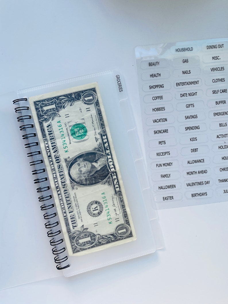 Peut inclure: Un classeur en plastique transparent contenant un billet d'un dollar. Le classeur est dot&eacute; d'une fermeture &agrave; glissi&egrave;re et d'une poche transparente sur le devant. La poche contient une vari&eacute;t&eacute; d'&eacute;tiquettes pour diff&eacute;rentes cat&eacute;gories, telles que "&eacute;picerie", "m&eacute;nage", "repas au restaurant", "beaut&eacute;", "essence", "divers", "sant&eacute;", "ongles", "v&eacute;hicules", "achats", "divertissements", "v&ecirc;tements", "caf&eacute;", "soir&eacute;e", "soins personnels", "loisirs", "cadeaux", "tampon", "vacances", "&eacute;pargne", "urgences", "soins de la peau", "d&eacute;penses", "factures", "animaux de compagnie", "enfants", "activit&eacute;s", "re&ccedil;us", "dettes", "vacances", "argent de poche", "allocation", "m&eacute;nage", "famille", "mois &agrave; venir", "No&euml;l", "Halloween", "Saint-Valentin", "Action de gr&acirc;ce", "P&acirc;ques", "anniversaires" et "juillet".
