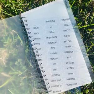 Peut inclure: Un classeur en plastique transparent avec une reliure spirale noire et des &eacute;tiquettes blanches pour la budg&eacute;tisation. Les &eacute;tiquettes comprennent des cat&eacute;gories telles que les &eacute;piceries, le m&eacute;nage, les sorties au restaurant, la beaut&eacute;, la sant&eacute;, les achats, le caf&eacute;, les loisirs, les vacances, les enfants, les dettes, l'allocation, le mois &agrave; venir, et plus encore.
