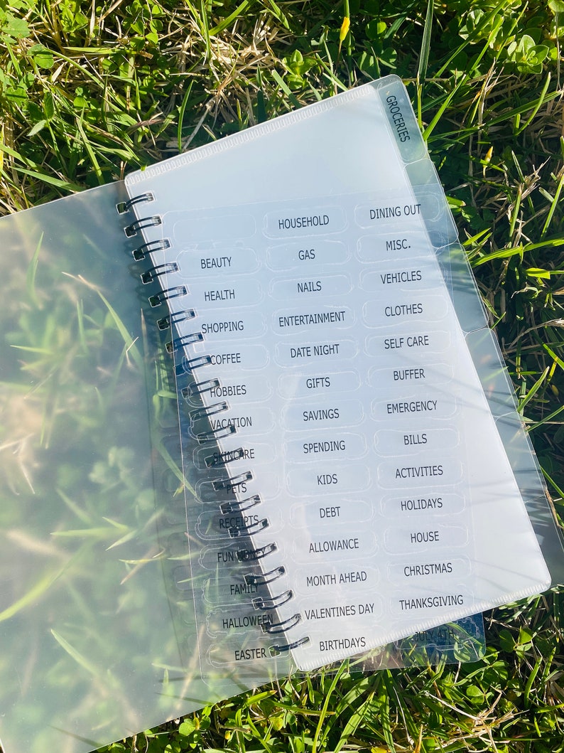 Peut inclure: Un classeur en plastique transparent avec une reliure spirale noire et des &eacute;tiquettes blanches pour la budg&eacute;tisation. Les &eacute;tiquettes comprennent des cat&eacute;gories telles que les &eacute;piceries, le m&eacute;nage, les sorties au restaurant, la beaut&eacute;, la sant&eacute;, les achats, le caf&eacute;, les loisirs, les vacances, les enfants, les dettes, l'allocation, le mois &agrave; venir, et plus encore.