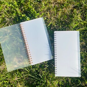 Peut inclure: Deux carnets &agrave; spirales en plastique transparent avec des anneaux dor&eacute;s. Les carnets sont pos&eacute;s sur un lit d'herbe verte.