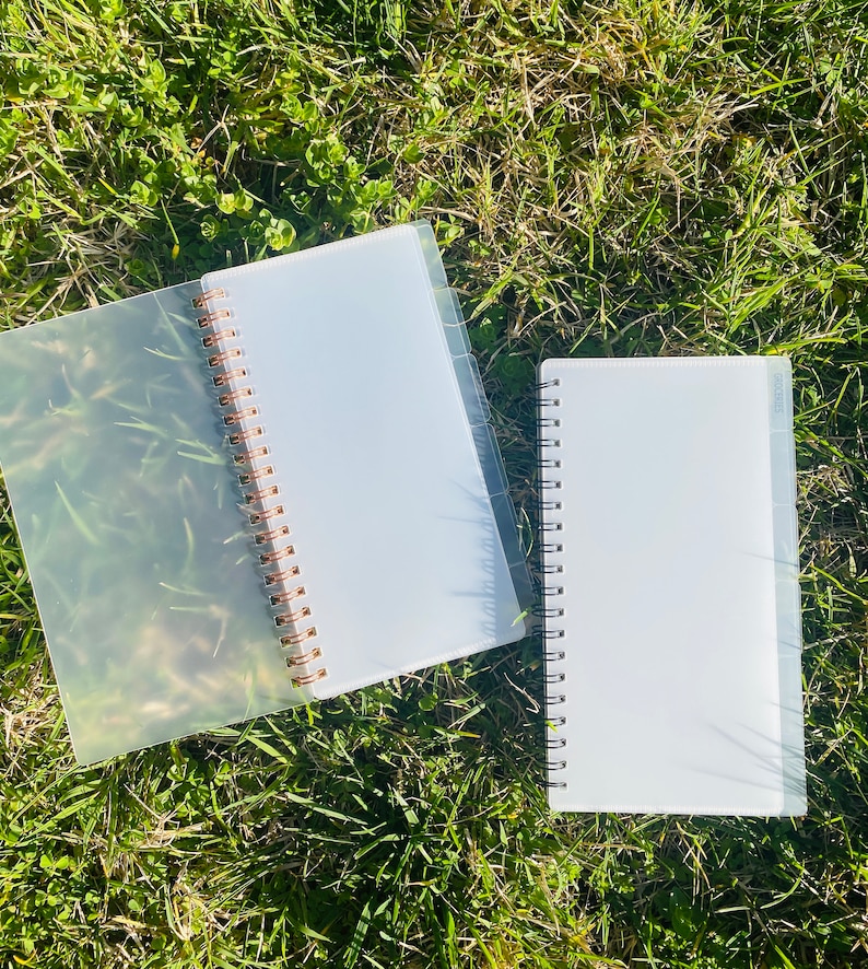 Peut inclure: Deux carnets &agrave; spirales en plastique transparent avec des anneaux dor&eacute;s. Les carnets sont pos&eacute;s sur un lit d'herbe verte.