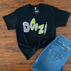 May include: Black t-shirt with a green and purple "Doozi!" graphic print. The shirt is laid out on a wooden surface with a pair of blue jeans.