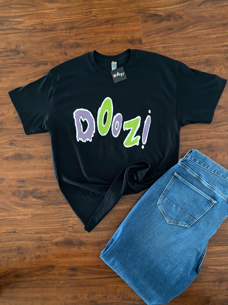 May include: Black t-shirt with a green and purple "Doozi!" graphic print. The shirt is laid out on a wooden surface with a pair of blue jeans.