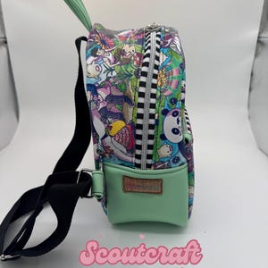 May include: A small, colorful backpack with a cartoon character print. The bag has a mint green front pocket and handle, a black strap, and a black and white striped zipper. The word "Scoutercraft" is written in pink script.