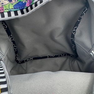 May include: Open view of a backpack interior. The bag has a gray lining and a black and white striped exterior. The top edge features a colorful cartoon design with characters and a zipper. The text "Scoutcraft" is visible.