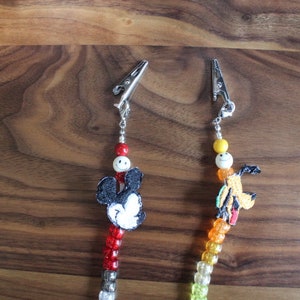 Op de afbeelding: Twee kralen fopspeenclips met metalen sluitingen. De ene clip heeft een Mickey Mouse-ontwerp en de andere een stripfiguur. De kralen zijn in verschillende kleuren, waaronder rood, wit, geel en oranje.