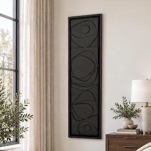 May include: A tall, rectangular wall art piece featuring a black frame and a matte black interior panel with raised, abstract geometric shapes including circles and curved lines arranged in a vertical composition on a white wall.