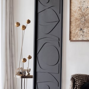 May include: A black framed wall art piece with a geometric design featuring abstract shapes and curves. The artwork is hanging on a white wall.