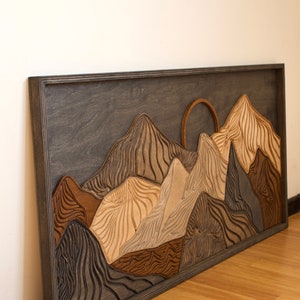 Puede incluir: Un cuadro rectangular de madera que representa un paisaje monta&ntilde;oso en capas. El dise&ntilde;o incluye picos tallados y texturizados en tonos marrones, beige, grises y carb&oacute;n, con un elemento solar circular sobre un fondo de color gris oscuro.