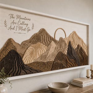 Puede incluir: Un cuadro rectangular de madera que muestra un paisaje monta&ntilde;oso en capas con tonos marrones claros, medios y oscuros. El texto "The Mountains Are Calling And I Must Go" est&aacute; impreso en negro en la parte superior izquierda del fondo de madera.