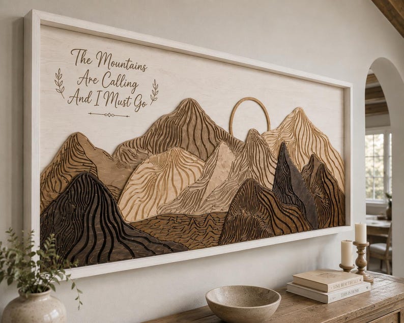 Puede incluir: Un cuadro rectangular de madera que muestra un paisaje monta&ntilde;oso en capas con tonos marrones claros, medios y oscuros. El texto "The Mountains Are Calling And I Must Go" est&aacute; impreso en negro en la parte superior izquierda del fondo de madera.