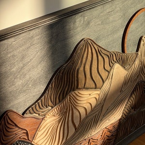 Puede incluir: Una pieza de arte mural de madera multicapa que presenta una cordillera estilizada. El dise&ntilde;o incorpora varios tonos de madera, incluyendo bronceado claro, marr&oacute;n medio y gris oscuro, con l&iacute;neas talladas que crean un efecto texturizado.