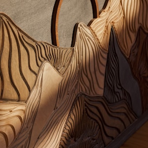 Puede incluir: Una pieza de arte mural de madera que presenta un paisaje monta&ntilde;oso tallado con texturas en capas y surcos profundos. Un anillo de madera circular se sit&uacute;a detr&aacute;s de los picos, creando una silueta similar a un sol sobre la madera.