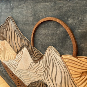 Puede incluir: Una pieza de arte mural de madera en capas con un fondo gris oscuro y cadenas monta&ntilde;osas talladas en tonos variados de beige claro, marr&oacute;n medio y marr&oacute;n oscuro. Un elemento solar arqueado de madera en tono &aacute;mbar sobre los picos.