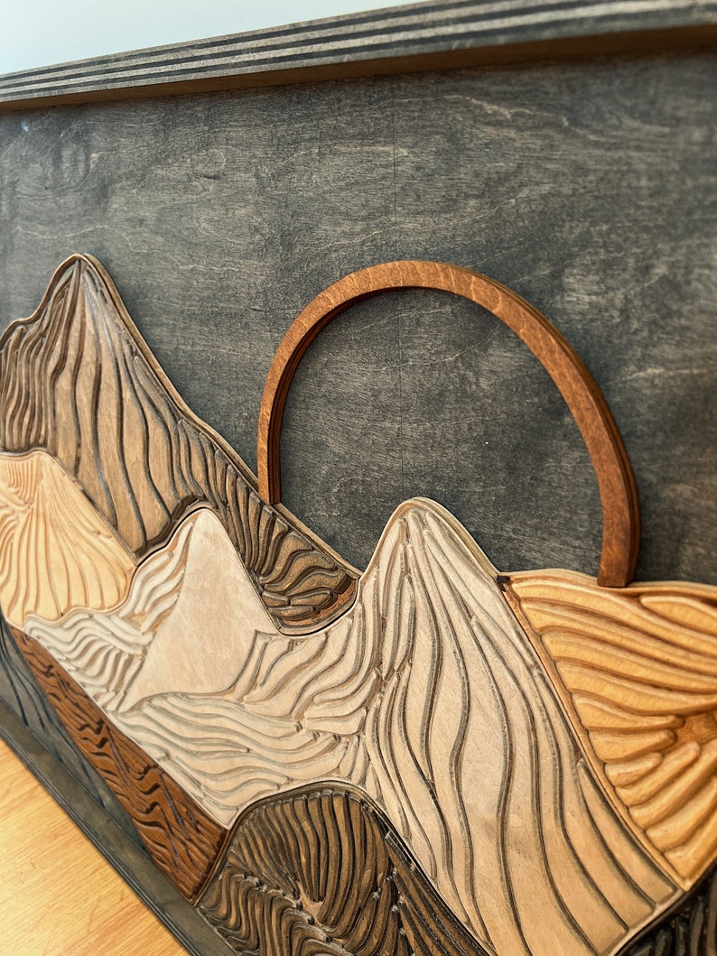 Puede incluir: Una pieza de arte mural de madera en capas con un fondo gris oscuro y cadenas monta&ntilde;osas talladas en tonos variados de beige claro, marr&oacute;n medio y marr&oacute;n oscuro. Un elemento solar arqueado de madera en tono &aacute;mbar sobre los picos.