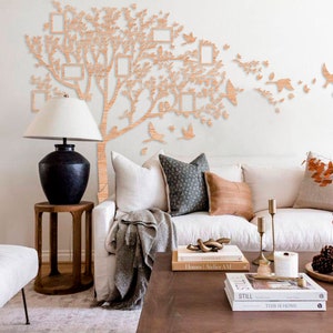 May include: A wooden family tree wall decoration featuring photo frames and bird motifs. The tree is mounted on a white wall above a white sofa with decorative cushions. A black lamp and wooden table are in the foreground.
