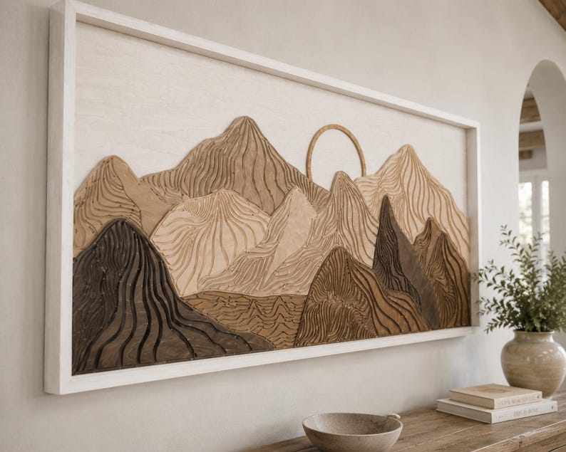 May include: A rectangular wall art piece featuring a layered wooden mountain landscape in shades of dark brown, light brown, and cream. The design includes a circular sun element and intricate carved line textures, all enclosed within a white frame.