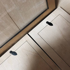 May include: Close-up of a wooden picture frame with a gold-coloured border and a clear glass pane. The frame is open, revealing the back of the frame and the wooden panels. Black hardware is visible.