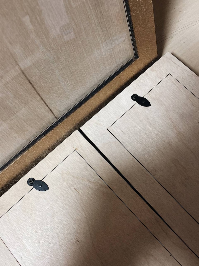 May include: Close-up of a wooden picture frame with a gold-coloured border and a clear glass pane. The frame is open, revealing the back of the frame and the wooden panels. Black hardware is visible.