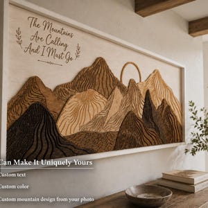 Puede incluir: Un cuadro rectangular de madera que muestra un paisaje monta&ntilde;oso en capas con diversos tonos de marr&oacute;n y beige. La obra incluye un sol tallado y el texto "The Mountains Are Calling And I Must Go" en una fuente cursiva oscura a la izquierda.