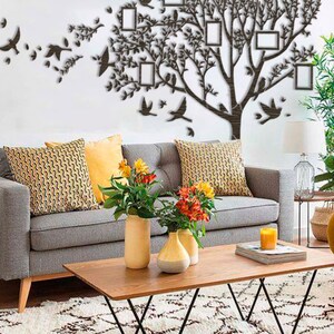 May include: A brown tree-shaped wall decoration with photo frames and bird silhouettes. The wall art is mounted on a white wall above a grey sofa with patterned cushions and a wooden coffee table. The room also features vases of flowers.