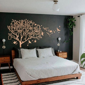 May include: A bedroom featuring a wooden bed frame, white bedding, and patterned pillows. A large tree wall decal with picture frames and birds adorns the dark wall. Two wooden bedside tables flank the bed, and a striped rug is on the floor.