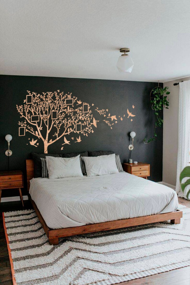 May include: A bedroom featuring a wooden bed frame, white bedding, and patterned pillows. A large tree wall decal with picture frames and birds adorns the dark wall. Two wooden bedside tables flank the bed, and a striped rug is on the floor.