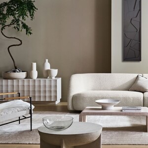 May include: A living room interior with a white sofa, a white chair, a coffee table with a glass bowl, and a wooden side table with a book and a white bowl. A large abstract wall art piece hangs on the wall behind the sofa. A potted bonsai tree sits on a white cabinet with a textured surface.