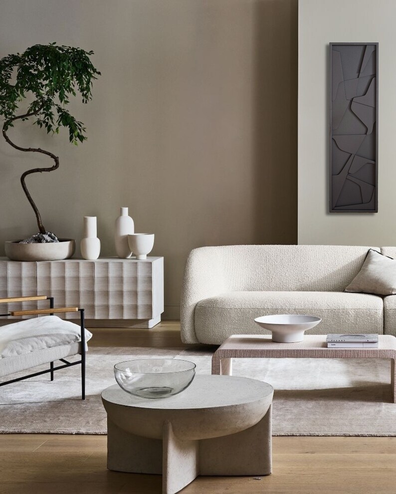 May include: A living room interior with a white sofa, a white chair, a coffee table with a glass bowl, and a wooden side table with a book and a white bowl. A large abstract wall art piece hangs on the wall behind the sofa. A potted bonsai tree sits on a white cabinet with a textured surface.