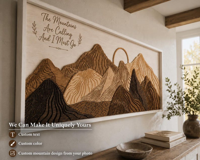 May include: A rectangular wooden wall art piece featuring a layered mountain landscape in various shades of brown and tan. The artwork includes a carved sun and the text "The Mountains Are Calling And I Must Go" in a dark script font on the left side.