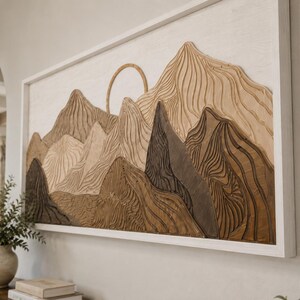 Puede incluir: Una pieza de arte mural rectangular que presenta un paisaje monta&ntilde;oso de madera en capas con un elemento solar circular. La obra muestra varios tonos de madera marr&oacute;n y bronceada con texturas onduladas talladas, todo dentro de un marco de madera blanco.