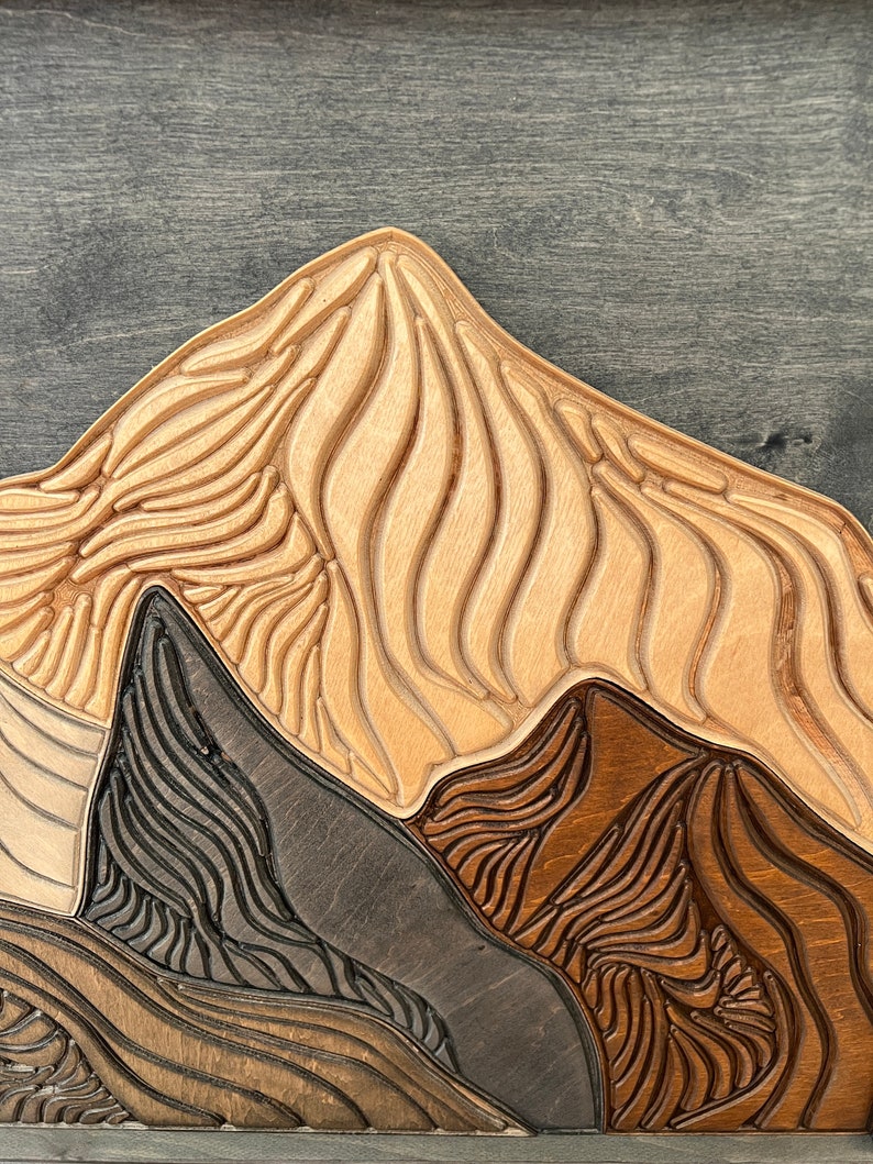Puede incluir: Una pieza de arte mural de madera en capas que presenta una cordillera estilizada. El dise&ntilde;o consiste en l&iacute;neas talladas de estilo topogr&aacute;fico en varios tonos de madera clara natural, marr&oacute;n medio y gris carb&oacute;n oscuro.