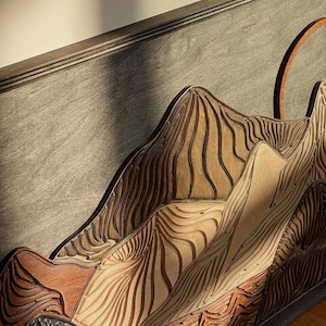Puede incluir: Un panel decorativo de madera para pared con un dise&ntilde;o de cordillera en capas. La pieza est&aacute; compuesta por varios tonos de madera, incluyendo beige claro, marr&oacute;n medio y gris oscuro, con l&iacute;neas verticales talladas y fluidas que imitan contornos.