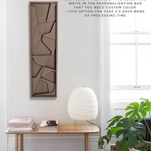 May include: A brown geometric wall art piece with a modern design. The art is hanging on a white wall above a wooden table with a white lamp and a green plant.