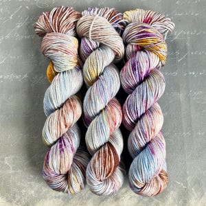 Könnte beinhalten: Drei Knäuel handgefärbtes Garn in einem Farbverlauf von Blau, Lila, Braun und Gelb.