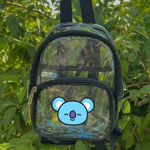 May include: Clear backpack with black trim and a small front pocket. The backpack features a blue koala graphic on the front pocket. The backpack has a top handle and adjustable shoulder straps. The background is green foliage.