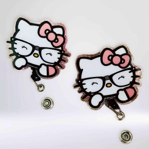 May include: Two Hello Kitty badge reels. Each reel features a white cat face with black glasses, a pink bow, and a pink and glittery border. The reels have a black retractable cord and a silver clip.
