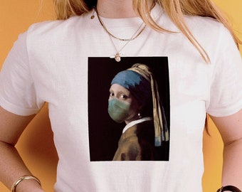May include: A white short-sleeved t-shirt featuring a graphic print of the Girl with a Pearl Earring wearing a green face mask. The artwork is centred on the chest. The shirt is paired with light blue denim jeans and a leopard print belt.