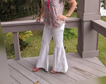 May include: A sleeveless collared shirt featuring a vibrant multi-colored floral pattern on a white background with a solid hot pink placket and collar, paired with white flared trousers and pink open-toe sandals with thin straps.