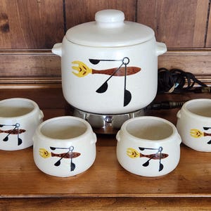 May include: A cream-colored ceramic electric fondue pot with a matching lid and four small matching bowls. Each piece features a graphic illustration of a yellow fork and black measuring spoons with a brown speckled handle design on the exterior.