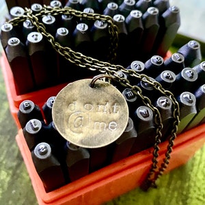 Pode incluir: Um colar de latão com um pingente redondo que diz "don't @ me". O colar está sobre uma caixa vermelha cheia de carimbos de letras de metal.